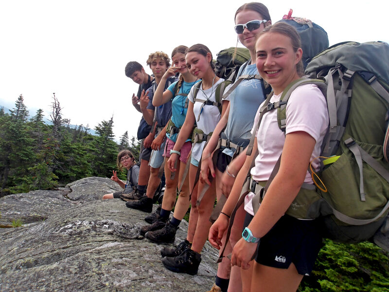 A group of young people are standing in a line on a rocky outcrop, likely during a hike. They are wearing backpacks and casual clothing, suggesting they are on an outdoor adventure. The background features trees and a somewhat cloudy sky. The overall impression is one of camaraderie and enjoyment of nature.